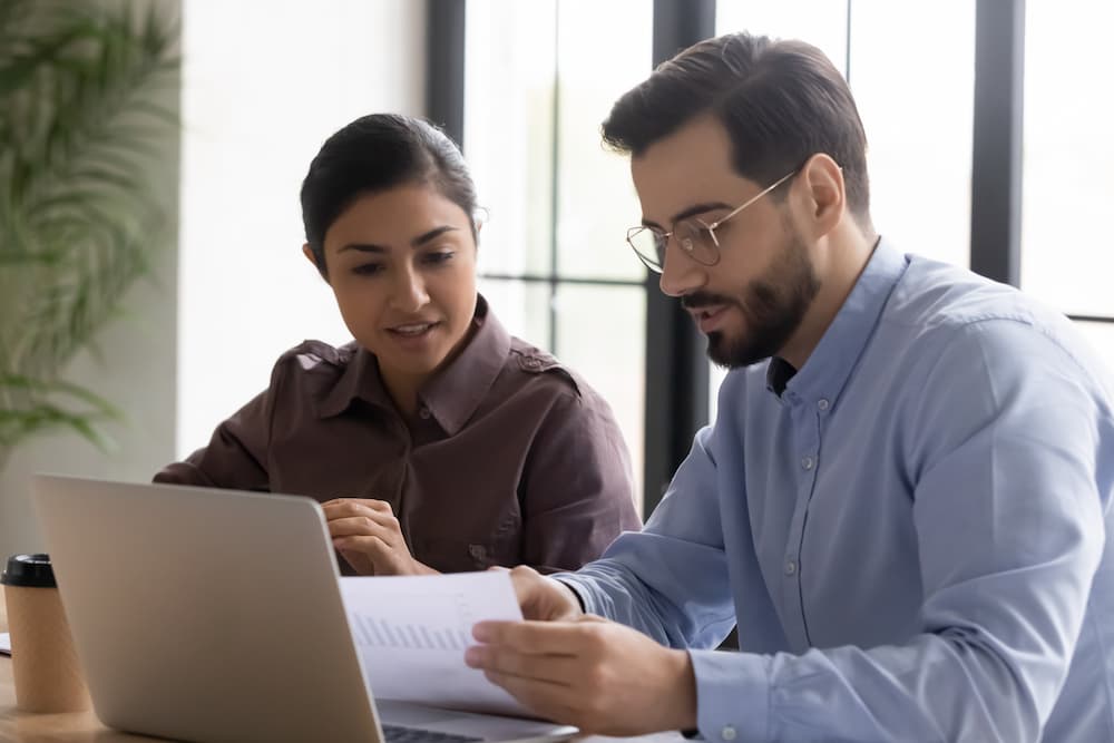 Ein Sales Manager sitzt mit einer Kollegin an einem Schreibtisch vor einem vor einem Laptop und bespricht ein Angebot für einen Kunden.
