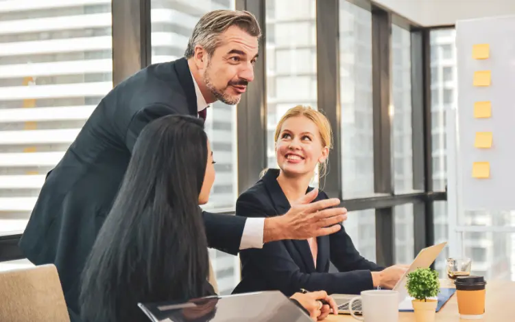 "Das Bild zeigt einen Head of Sales mit einem freundlich, dynamischen Gesichtsausdruck, der leicht nach vorn gebeugt zwischen zwei sitzenden Sales Managerinnen an einem Konferenztisch steht und seine Teamkolleginnen bei einer Vertragsverhandlung unterstützt."