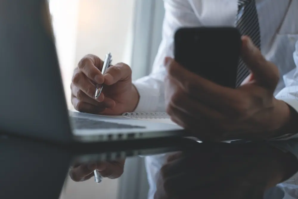 Account Executive Symbolbild: Ein Mann sitzt vor einem Laptop. In der linken Hand hält er ein Smartphone und macht sich mit der rechten Hand eine Notiz auf einem Schreibblock.
