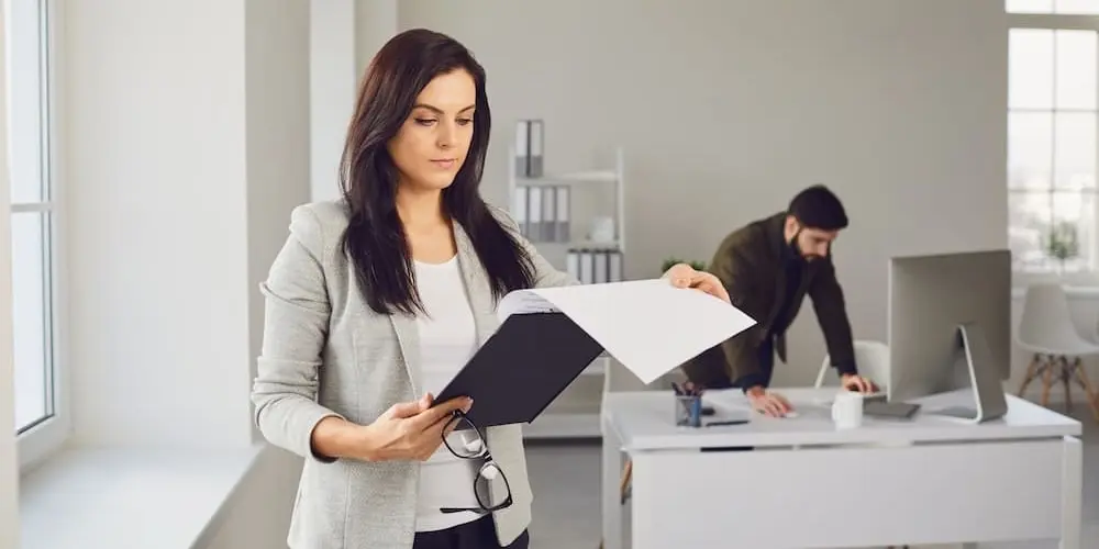 Im Vordergrund ist eine Vertriebscontrollerin zu sehen, die sich Unterlagen auf einem Clipboard anschaut. Im Hintergrund beugt sich ein Kollege an einem Schreibtsich vor und betrachtet einen PC-Bildschirm.