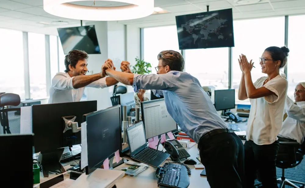 Vertriebsassistent Symbolbild: Ein Vertriebsassistent freut sich gemeinsam mit dem Vertriebsteam über einen Verkaufserfolg. Zu sehen ist eine Gruppe aus fünf Personen in einer Office-Umgebung. Zwei Personen im Vordergrund reichen sich über den Schreibtisch hinweg die Hände.