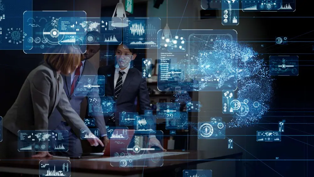 IT-Vertriebler Symbolbild: Das Bild zeigt im Vordergrund mehrere digitale Grafiken. Im Hintergrund sind drei Personen in Businesskleidung zu sehen, die stehendan einem Tisch um einen Laptop versammelt sind.