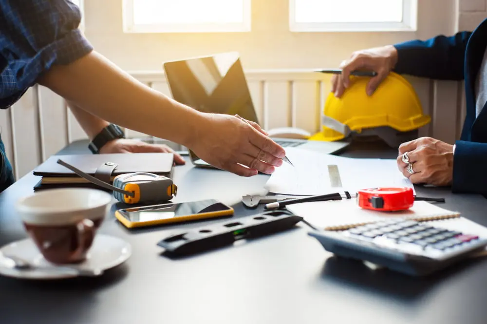 "Technischer Vertrieb Symbolbild: Ein technischer Vertriebsmitarbeiter bespricht ein Projekt mit einem Bauleiter. Zu sehen sind die Arme zweier Menschen, die an einem Schreibtisch arbeiten. Auf dem Schreibtisch befinden sich Taschenrechner, Smartphone, Laptop und Konstruktionspläne."